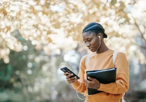 A student looking at their phone with airpods in, holding some files